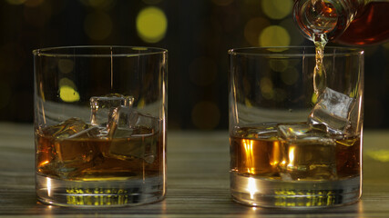 Pouring of golden whiskey, cognac or brandy from bottle into glass with ice cubes. Shiny background