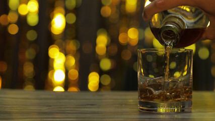 Pouring of golden whiskey, cognac or brandy from bottle into glass with ice cubes. Shiny background