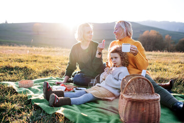 Small girl with mother and grandmother having picnic in nature at sunset.