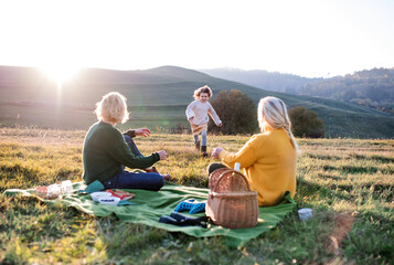 Small girl with mother and grandmother having picnic in nature at sunset.