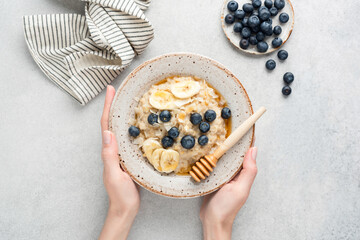 Female hands holding bowl of oatmeal porridge with banana slices, blueberries, coconut flakes and...