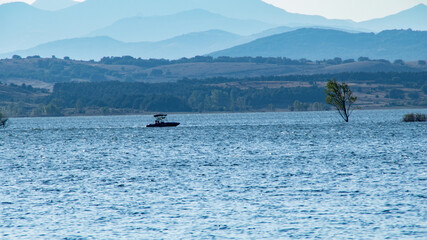 Obraz premium Pequeño barco navegando en un tranquilo lago azul en una tarde de verano con unas difuminadas montañas azules en el fondo.