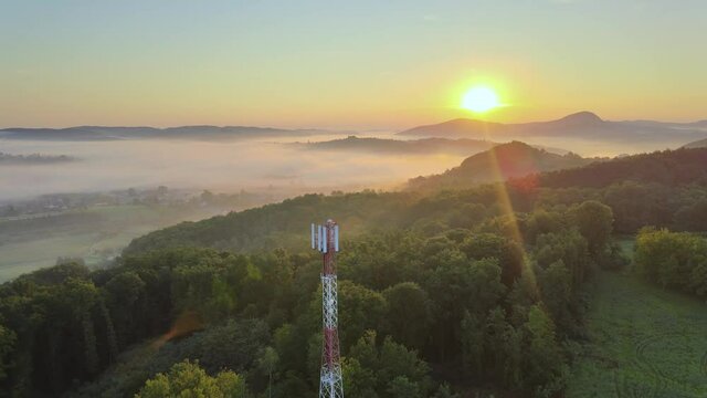 Isolated 5G/4G telecommunication tower standing tall on green mountains with sun shining in the background at golden hour. Backward moving aerial view.