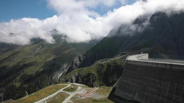 Large dam in Switzerland in the middle of the mountain with some rolling clouds over green fields.