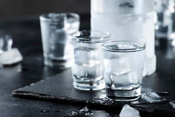 Vodka in shot glasses on black stone background, iced strong drink in misted glass