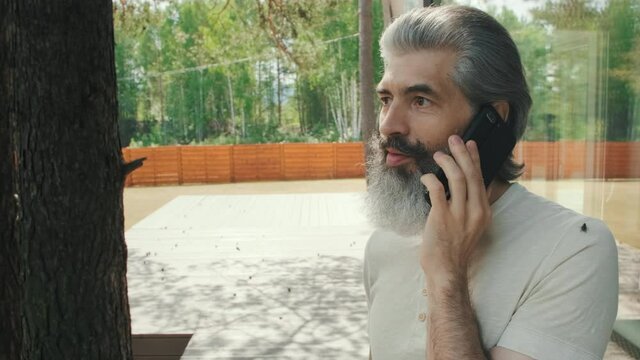 Tracking shot of stylish senior man with grey beard and moustache standing on porch of house with panoramic windows and talking on phone