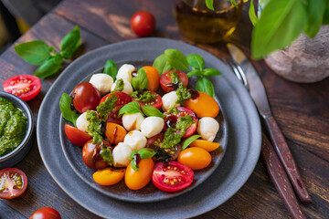 Caprese salad with fresh cherry tomatoes, mini mozzarella, pesto, basil, olive oil and balsamic sauce on blue plate, wooden rustic table. Mediterranean diet concept. Italian healthy food. Copy space.