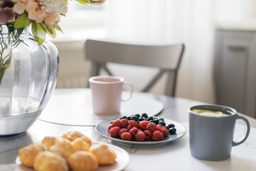 Beautiful breakfast on table near window in white kitchen - cups of tea, flowers, berries, snacks. Healthy, romantic meal in modern room interior. Stylish room interior with natural morning light.
