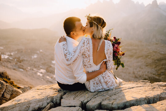 Beautiful bride and bridegroom kissing and hugs sitting on stone cliff at sunset