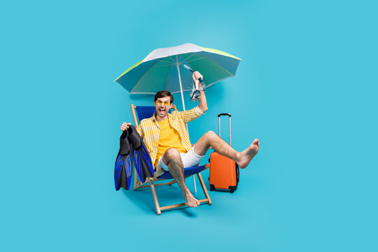 Full length photo crazy guy sit lounger umbrella rest relax swim bottom coral reef hold flippers goggles luggage baggage bag wear yellow white shirt shorts isolated blue color background