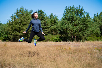 Young woman in sports outfit hops high on a natural meadow with trees and blue sky in the background