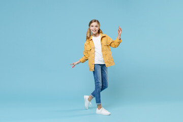 Full length portrait of funny little blonde kid girl 12-13 years old in yellow jacket isolated on blue background. Childhood lifestyle concept. Mock up copy space. Pointing index finger up, dancing.