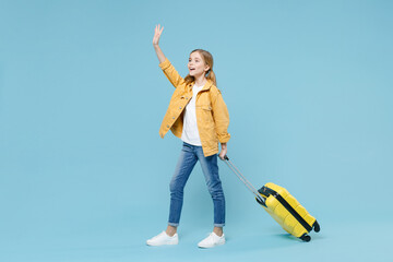 Full length portrait of little kid girl 12-13 years old isolated on blue background. Passenger traveling abroad on weekends. Air flight journey concept Hold suitcase waving greet with hand catch taxi.