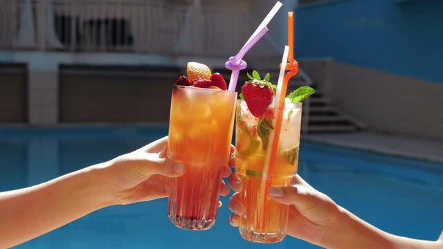 Close up view of young female hands holding exotic non-alcoholik ice fruit cocktails, making cheer, clink glasses, joy drinking and celebrate friendly meeting near pool in luxury spa complex.