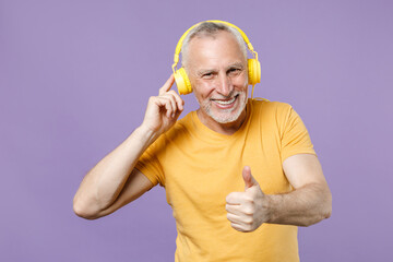 Smiling elderly gray-haired mustache bearded man in casual yellow t-shirt posing isolated on violet background studio portrait. People lifestyle concept. Listen music with headphones showing thumb up.