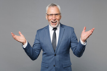 Excited elderly gray-haired bearded business man in classic blue suit shirt tie posing isolated on grey background . Achievement career wealth business concept. Keeping mouth open, spreading hands.