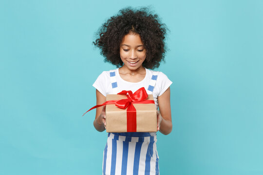 Excited little african american kid girl 12-13 years old in striped clothes isolated on blue background. International Women's Day birthday, holiday concept. Hold red present box with gift ribbon bow.