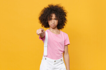 Dissatisfied little african american kid girl 12-13 years old in pink t-shirt isolated on yellow background in studio. Childhood lifestyle concept. Mock up copy space. Pointing index finger on camera.