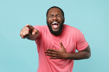Fototapeta premium Laughing young african american man guy in casual pink t-shirt posing isolated on blue background. People lifestyle concept. Mock up copy space. Pointing index finger on camera, put hand on stomach.