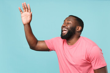 Cheerful young african american man guy in casual pink t-shirt posing isolated on blue background studio. People lifestyle concept. Mock up copy space. Waving greeting with hand as notices someone.