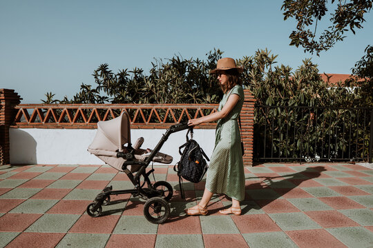 Stylish mother in hat walking with stroller on exotic street