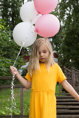 A beautiful blonde girl of six seven years old with balloons, in bright yellow clothes. The girl celebrates her birthday in a summer park.
