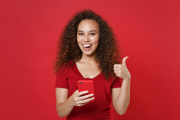 Funny young african american girl in casual t-shirt posing isolated on red wall background studio portrait. People lifestyle concept. Mock up copy space. Using mobile cell phone, showing thumb up.