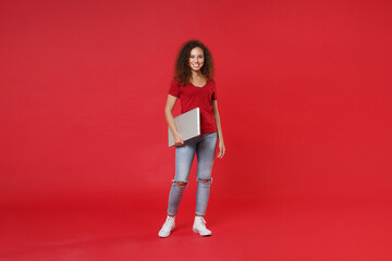 Full length portrait of smiling young african american girl in casual t-shirt isolated on red wall background studio portrait. People lifestyle concept. Mock up copy space. Hold laptop pc computer.