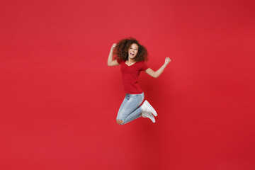 Full length portrait of joyful young african american girl in casual t-shirt isolated on red background studio portrait. People lifestyle concept. Mock up copy space. Jumping doing winner gesture.