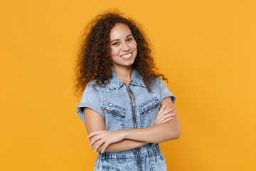 Smiling young african american woman girl in casual denim clothes isolated on yellow background studio portrait. People sincere emotions lifestyle concept. Mock up copy space. Holding hands crossed.