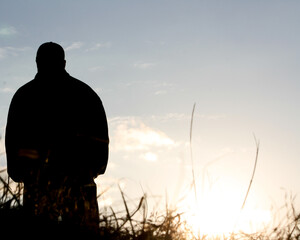 Silhouette of a man standing on the left side of the frame looking out at the sunset from a field of grass