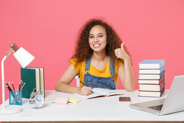 Smiling young african american girl employee in office sit work at desk isolated on pink wall...