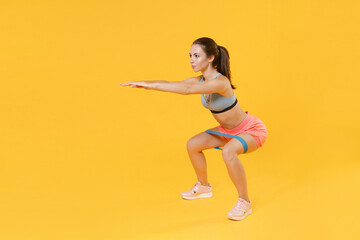Full length portrait of young fitness sporty woman girl in sportswear working out isolated on yellow background. Workout sport motivation lifestyle concept. Doing exercise squatting with fitness gums.