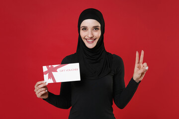 Smiling young arabian muslim woman in hijab black clothes posing isolated on bright red background. People religious lifestyle concept. Mock up copy space. Hold gift certificate, showing victory sign.