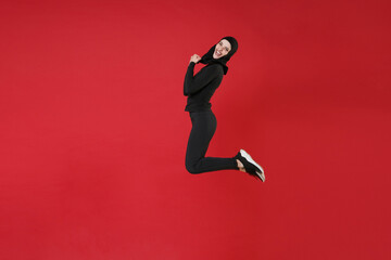 Fototapeta na wymiar Full length portrait side view of happy young arabian muslim woman in hijab black clothes posing isolated on red background studio. People religious lifestyle concept. Jumping, doing winner gesture.