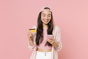 Smiling young asian woman girl in casual clothes cap posing isolated on pastel pink wall background studio. People lifestyle concept. Mock up copy space. Using mobile cell phone hold credit bank card.