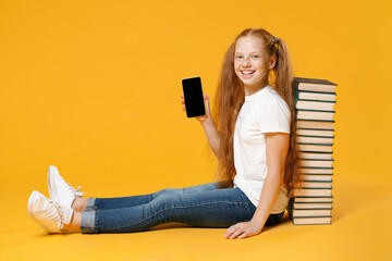 Full length redhead young girl 12-13 years old in white tshirt sit near big stack school book hold using mobile cell phone isolated on yellow background children studio portrait Kids education concept