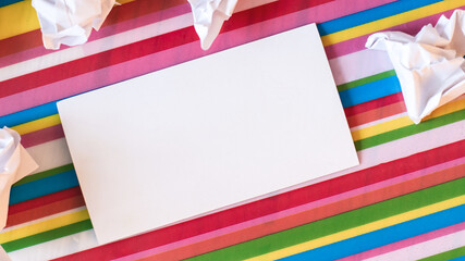 Fototapeta premium A white blank card lies on a multi-colored bright striped background near crumpled pieces of paper. Place to insert text. Close-up. Business concept