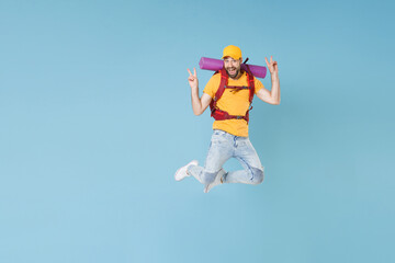 Full length portrait Excited young traveler man in cap with backpack isolated on blue background. Tourist traveling on weekend getaway. Tourism discovering hiking concept. Jump showing victory sign.