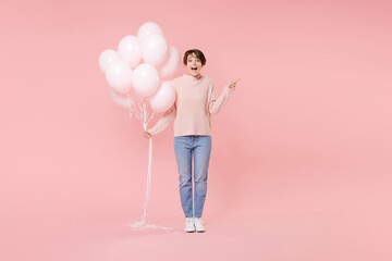 Full length portrait of shocked young girl in casual sweater isolated on pink background. Birthday holiday party concept. Mock up copy space. Celebrating hold air balloons pointing index finger aside.