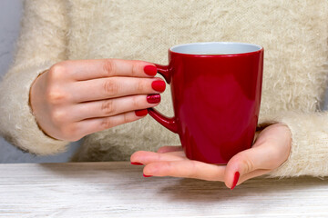 Female hands hold a red cup against the background of a white fluffy sweater with a beautiful red winter manicure. Drink, fashion, morning. New Year. Celebration. Tea coffee.
