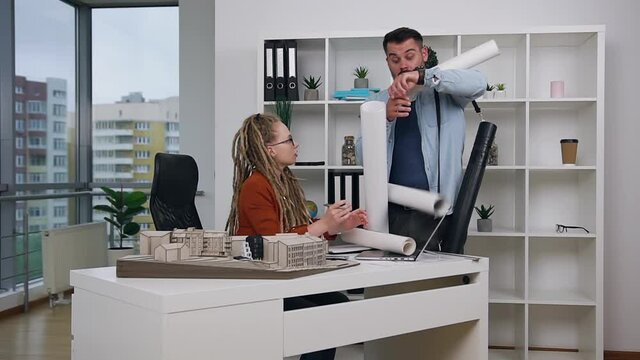 Likable funny bearded young architect carrying different blueprints to his strict concentrated successful modern female chief and unexpectedly droping them on her workplace