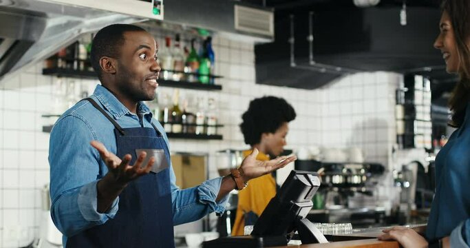 Cheerful African American waiter serving coffee and talking with Caucasian woman at counter in cafe. Man barrista having nice talk with customer. Male bartender chatting with female client Multiethnic