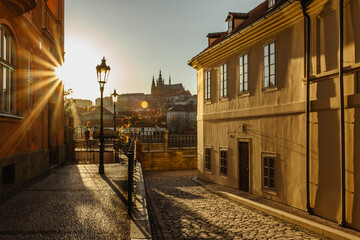 Beautiful view of a cobblestone pedestrian street and Prague Castle with sun rays. Tranquil city scene. Walking through the empty capital of Czechia. Prague panorama at sunset. Evening sunlight.