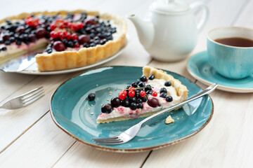 Blueberry open pie on blue plate with cup of black tea and white tea pot on bright painted wood background
