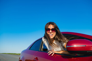 Smiling young female stick out from vehicle while travel, wearing sunglasses at sky and empty road background. The girl looks into the camera.