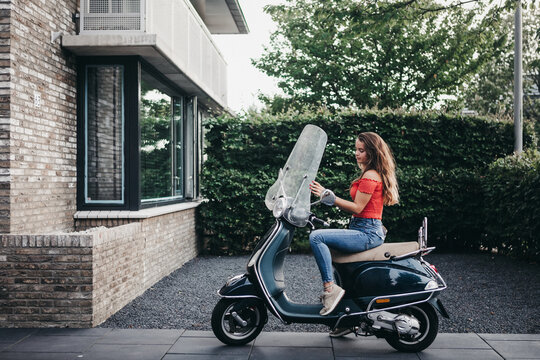 Pretty teen with long hair sitting on a vespa scooter in front of a house