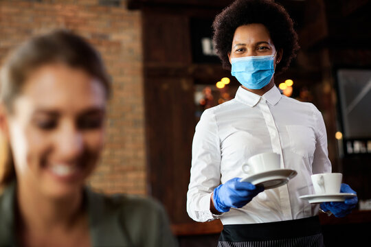 Happy Black Waitress With Protective Face Mask And Gloves Serving Coffee In A Cafe.