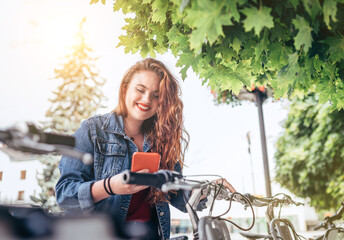 Fototapeta na wymiar Portrait of smiling red curled long hair caucasian teen girl block out bike at Bicycle sharing point using the modern smartphone. Green urban transport and modern technology devices concept image.