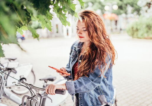 Portrait of smiling red curled long hair caucasian teen girl unlocking bike at Bicycle sharing point using the modern smartphone. Green urban transport and modern technology devices concept image.